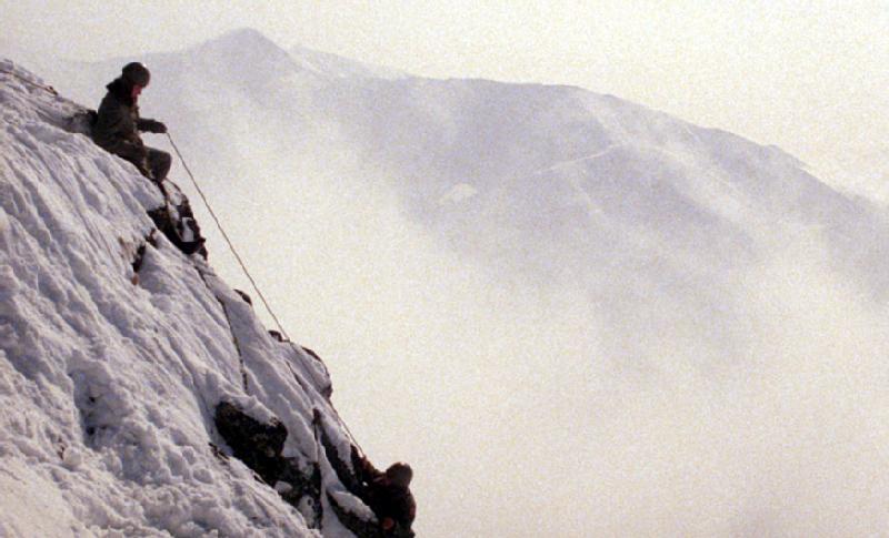Dwóch wspinaczy wspina się na strome, pokryte śniegiem zbocze góry, asekurując się linami. W tle widać zamglone, ośnieżone szczyty.