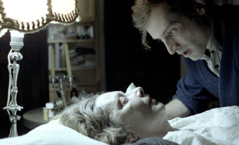 Młody mężczyzna pochyla się nad kobietą leżącą w łóżku pod kocem, oświetloną starą lampą. Scena ma ponury, intymny nastrój, mężczyzna patrzy na kobietę, która wydaje się być chora lub śpiąca.