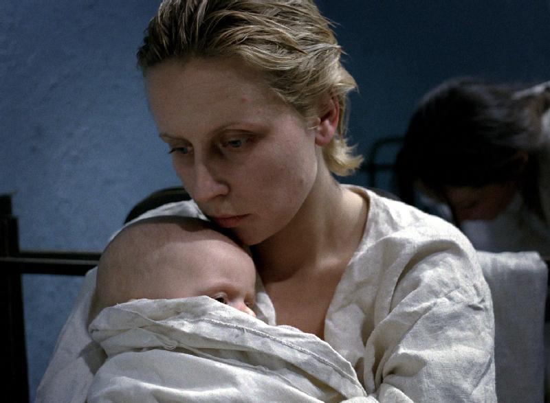 Kobieta z krótkimi blond włosami, ubrana na biało, trzyma dziecko blisko piersi. Wygląda na ponurą i zamyśloną. Inna osoba jest rozmazana w tle na tle zwykłej niebieskiej ściany.