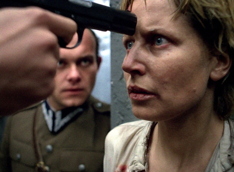 Zrozpaczona kobieta o krótkich blond włosach staje twarzą w twarz z pistoletem wymierzonym w jej czoło, podczas gdy za nią stoi surowy mężczyzna w mundurze wojskowym, uważnie się przyglądając. Scena jest napięta i dramatyczna.