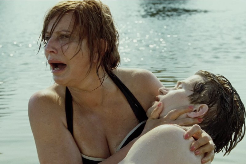 Zrozpaczona kobieta w kostiumie kąpielowym trzyma nieprzytomnego młodego mężczyznę w płytkiej wodzie. Na jej twarzy widać strach i poczucie pilności.