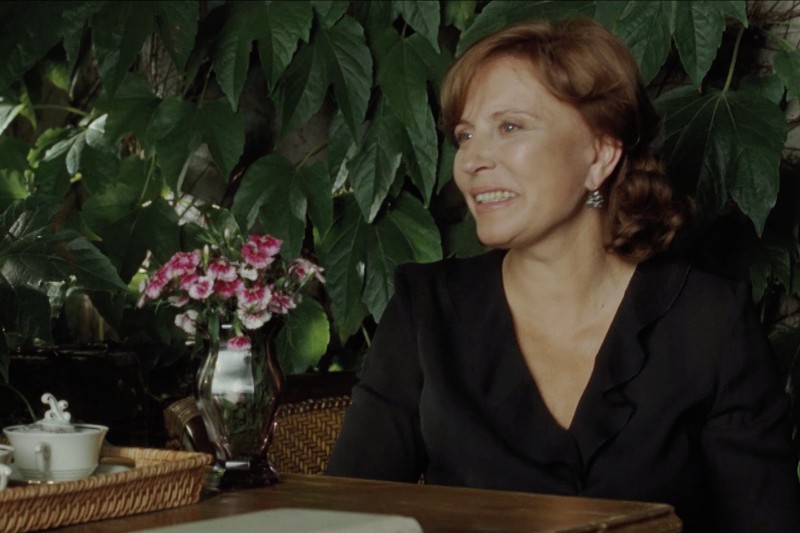 Kobieta o jasnobrązowych włosach, ubrana w czarny top, siedzi przy drewnianym stole z małym wazonem z różowymi kwiatami i filiżanką, uśmiechając się. Bujne zielone liście tworzą tło.