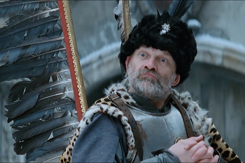 Mężczyzna ubrany w średniowieczną zbroję i futrzaną czapkę z piórkową peleryną stoi ze splecionymi palcami, patrząc w górę z poważnym wyrazem twarzy na tle ośnieżonego, kamiennego tła.