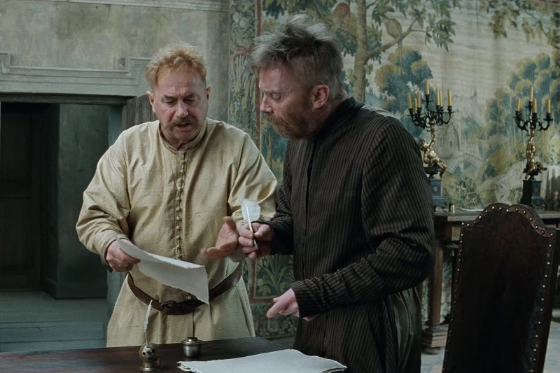 Dwóch mężczyzn w staromodnych ubraniach stoi przy drewnianym stole, trzymając papiery i pióro. Za nimi znajduje się dekoracyjny gobelin ścienny i świecznik, sugerujący historyczne lub średniowieczne otoczenie.