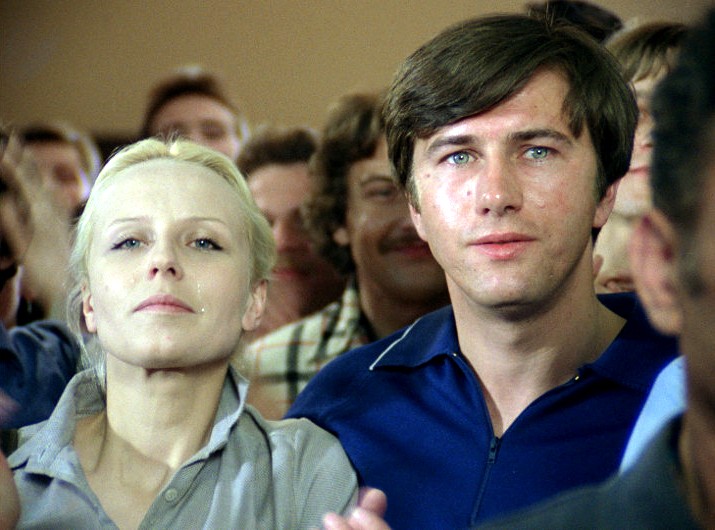 Blondynka i ciemnowłosy mężczyzna stoją blisko siebie w tłumie, oboje patrzą przed siebie z poważnymi minami. Ludzie w tle są lekko rozmazani.