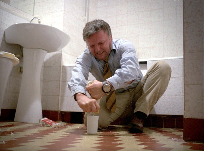 Mężczyzna w koszuli i krawacie kuca na podłodze w łazience, krzywiąc się, gdy wyciska torebkę herbaty do kubka. Na kafelkowej podłodze widać rozlane przedmioty i wodę.