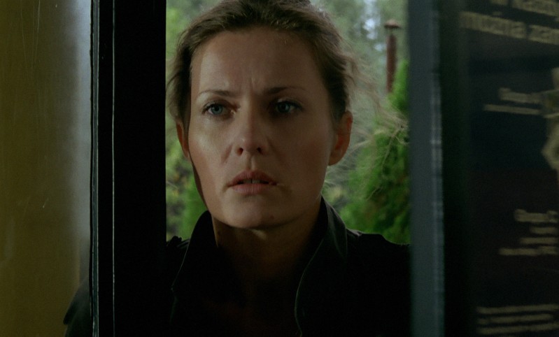 Kobieta o jasnobrązowych włosach i poważnym wyrazie twarzy stoi częściowo w drzwiach, patrząc przed siebie. Na rozmytym tle na zewnątrz widać bujną zieleń.