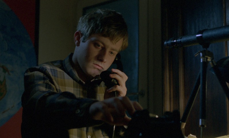 Młody mężczyzna w kraciastej koszuli rozmawia przez telefon z tarczą w słabo oświetlonym pokoju, sięgając po telefon jedną ręką. W tle widać teleskop i częściowo widoczną mapę świata.