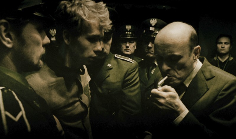 Napięta scena, w której kilku mężczyzn w mundurach wojskowych otacza młodego mężczyznę, podczas gdy łysy mężczyzna w garniturze zapala papierosa. Sceneria wydaje się mroczna i intensywna.