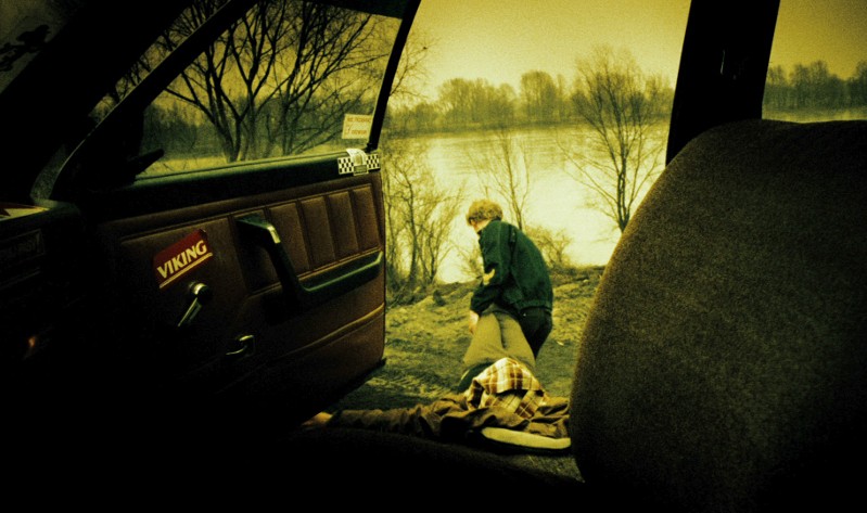 Osoba stoi na zewnątrz przy brzegu rzeki, widziana przez otwarte drzwi samochodu. Wnętrze samochodu jest częściowo widoczne, a scena ma żółtozielony odcień, tworząc nastrojowy, atmosferyczny efekt. Nagie drzewa wyznaczają tło.