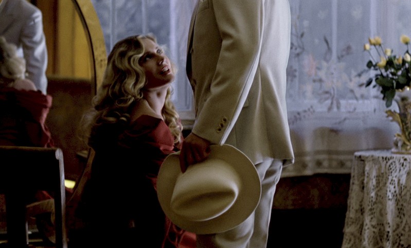 Kobieta w czerwonej sukience klęczy obok stojącego mężczyzny w białym garniturze, który trzyma biały kapelusz. Pokój jest delikatnie oświetlony koronkowymi firankami i wazonem z żółtymi kwiatami na pokrytym koronką stole.