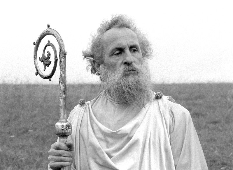 Starszy mężczyzna z długą brodą i kręconymi włosami, ubrany w powłóczystą białą szatę, trzyma ozdobny kij ze spiralnym wzorem. Stoi na zewnątrz na trawiastym polu, zamyślony, patrząc w bok.