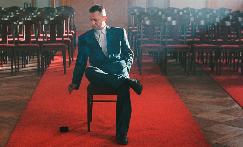 Mężczyzna w garniturze siedzi samotnie na krześle pośrodku czerwonego dywanu, otoczony pustymi drewnianymi krzesłami, z ręką wyciągniętą w kierunku małego, czarnego przedmiotu leżącego na podłodze.