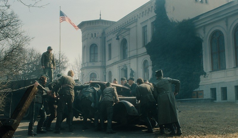 Grupa żołnierzy w długich płaszczach pcha zabytkowy samochód przed dużym, starym budynkiem z amerykańską flagą powiewającą na maszcie. Nagie drzewa i zimowe światło sugerują zimną porę roku.