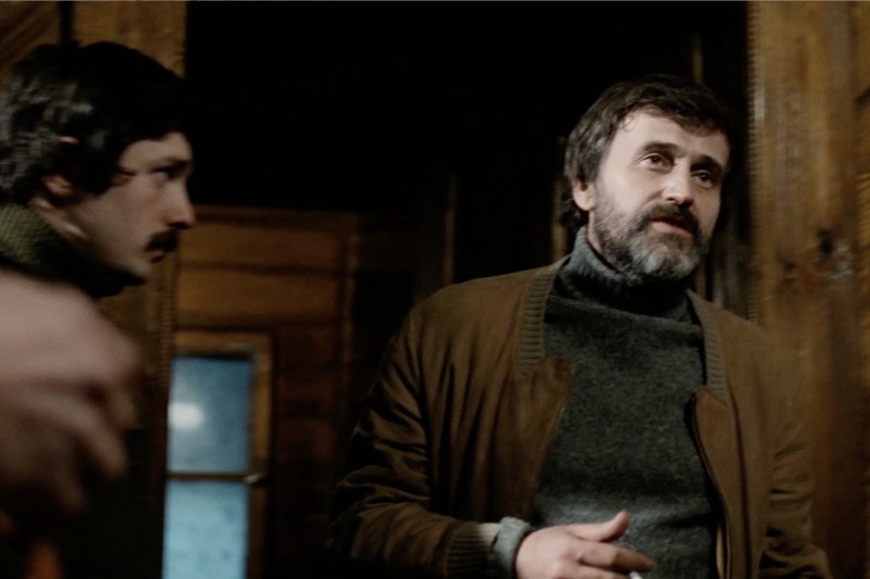 Dwóch mężczyzn w ciemnych kurtkach i golfach stoi w słabo oświetlonym drewnianym pokoju. Jeden mężczyzna z brodą jest lekko zwrócony twarzą w stronę kamery, podczas gdy drugi stoi w tle, częściowo w cieniu.