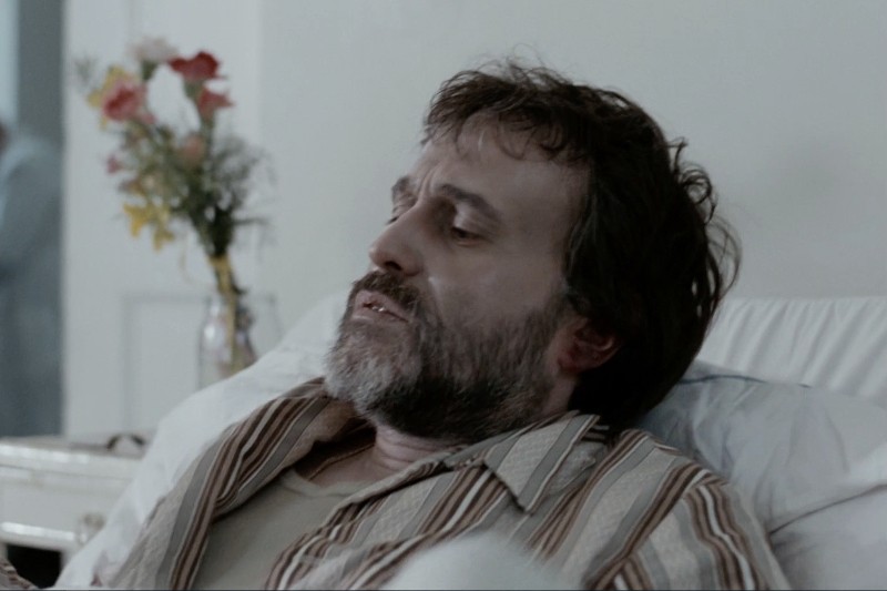Brodaty mężczyzna z rozczochranymi włosami leży na szpitalnym łóżku, wygląda na chorego i zamyślonego. Wazon z kwiatami stoi na stole w tle, na tle prostej, jasnej ściany.