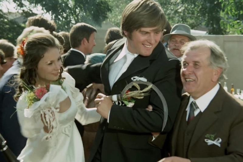 Panna młoda i pan młody w strojach ślubnych uśmiechają się i trzymają za ręce, świętując w towarzystwie starszego mężczyzny w garniturze wśród tłumu na zewnątrz.