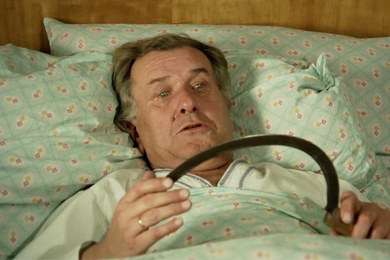 Starszy mężczyzna w piżamie leży w łóżku pod wzorzystym kocem, patrzy w górę ze zdziwionym wyrazem twarzy i trzyma sierp w rękach.