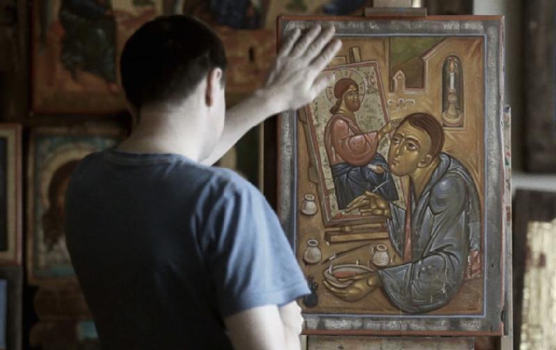 Osoba w niebieskiej koszuli delikatnie dotyka ikony religijnej przedstawiającej artystę malującego świętego, w tle widoczne są inne podobne ikony.