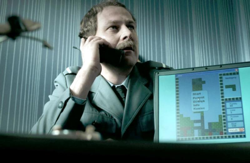 Mężczyzna w zielonym mundurze wojskowym rozmawia przez telefon, siedząc przy biurku. Przed nim znajduje się ekran komputera wyświetlający grę Tetris. W tle widać niebieską, pasiastą ścianę.