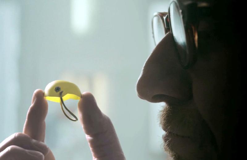 Osoba nosząca okulary uważnie przygląda się małemu żółtemu przedmiotowi w kształcie półkuli z czarną pętlą, trzymając go delikatnie między palcami.