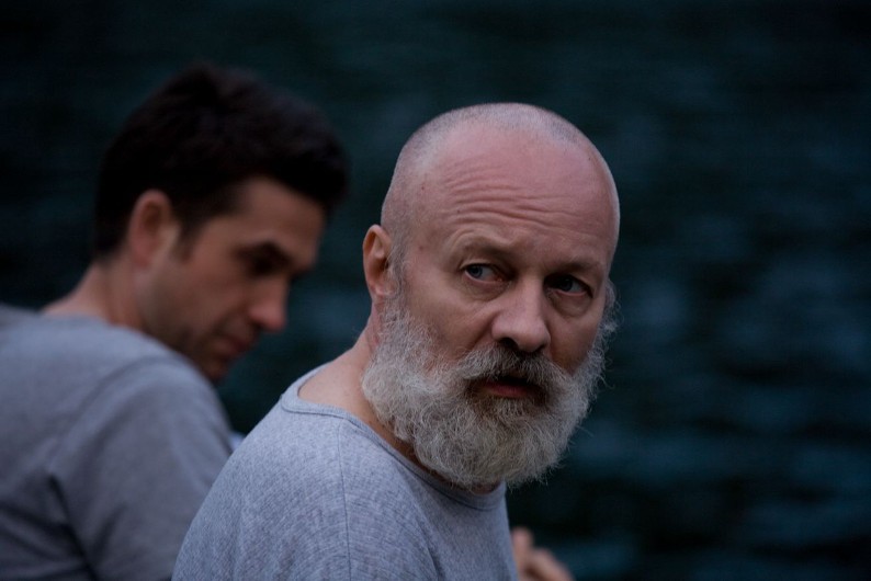 Brodaty mężczyzna z poważnym wyrazem twarzy spogląda lekko przez ramię, podczas gdy inny mężczyzna z krótkimi, ciemnymi włosami siedzi rozmazany w tle. Obaj mają na sobie szare koszule, a za nimi znajduje się ciemne, nieostre tło.