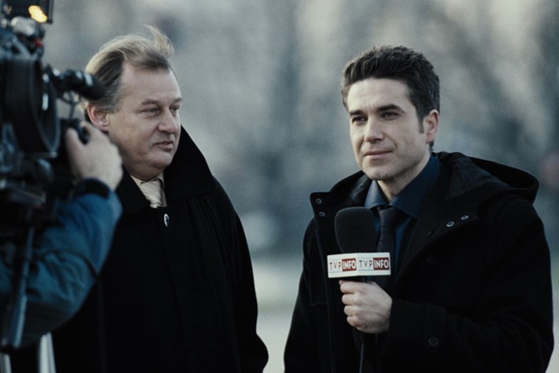 Reporter trzymający mikrofon przeprowadza wywiad z mężczyzną w płaszczu na zewnątrz, podczas gdy jest filmowany przez kamerzystę. Obaj mężczyźni mają poważne miny i są formalnie ubrani. Tło jest rozmyte.