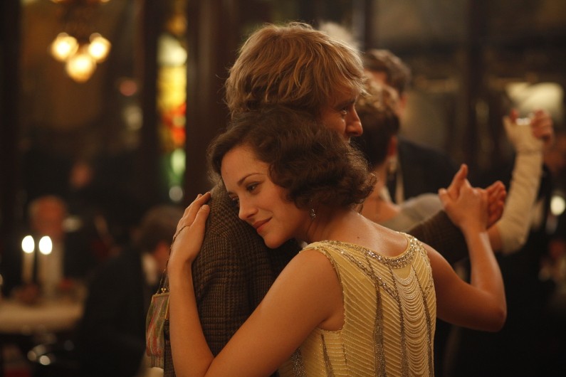 Kobieta w koralikowej sukni delikatnie obejmuje mężczyznę, wykonując powolny taniec w ciepło oświetlonej, eleganckiej sali balowej, otoczona innymi tancerzami w formalnych strojach.