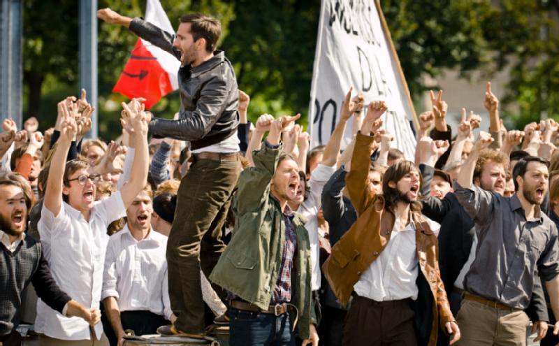 Duża grupa ludzi stoi na zewnątrz, podnosząc pięści i krzycząc na znak protestu. Niektórzy trzymają transparenty i flagi, a ich miny pokazują silne emocje i determinację. W tle widać drzewa.