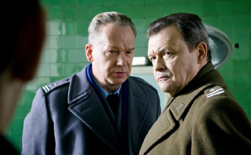 Dwóch mężczyzn w mundurach wojskowych stoi blisko siebie przed zieloną ścianą z kafelków, patrząc poważnie na kogoś poza kadrem. Ich wyrazy twarzy są napięte i skupione.