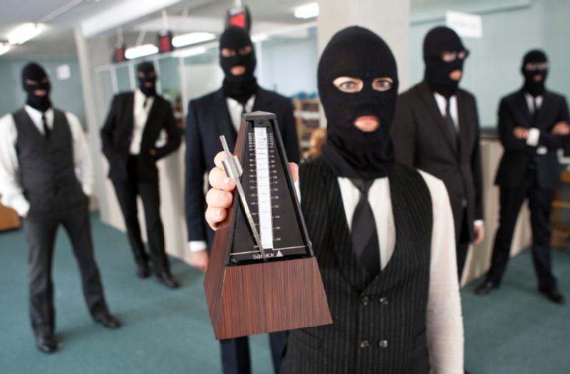 Sześć osób w garniturach i czarnych maskach narciarskich stoi w biurze. Osoba z przodu trzyma drewniany metronom w stronę kamery, podczas gdy inni stoją w tle, ze skrzyżowanymi ramionami lub rękami na biodrach.