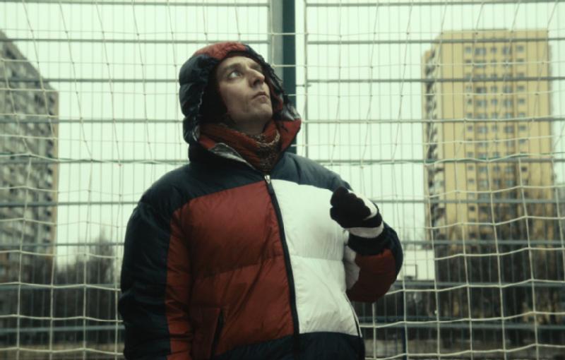 Osoba w czerwonej, czarnej i białej kurtce puchowej i kapturze stoi na zewnątrz przed metalowym płotem, w tle widać budynki mieszkalne. Niebo jest zachmurzone.