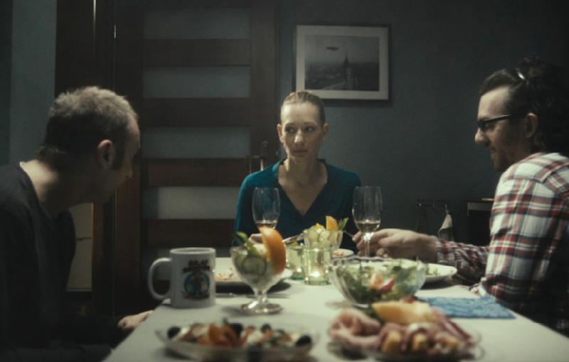Trzy osoby siedzą przy stole jadalnym zastawionym jedzeniem, owocami i kieliszkami do wina. Kobieta w środku wygląda poważnie, podczas gdy dwaj mężczyźni po obu stronach są zajęci rozmową. Scena jest słabo oświetlona.