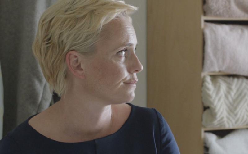 Osoba z krótkimi blond włosami i granatowym topem patrzy w prawo. W tle, złożone ręczniki lub koce są ułożone na drewnianych półkach.