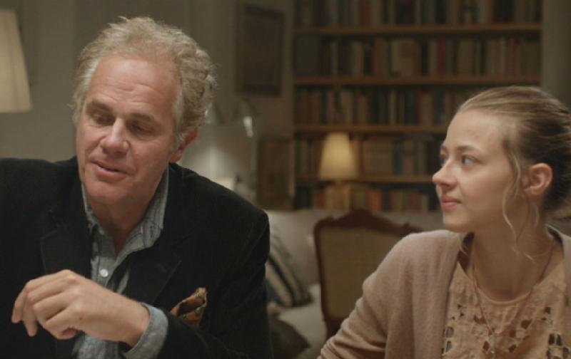 Mężczyzna w średnim wieku z siwymi włosami i młodsza kobieta z blond włosami siedzą razem przy stole w ciepło oświetlonym pokoju z regałami w tle. Mężczyzna patrzy przed siebie, podczas gdy kobieta patrzy na niego i lekko się uśmiecha.