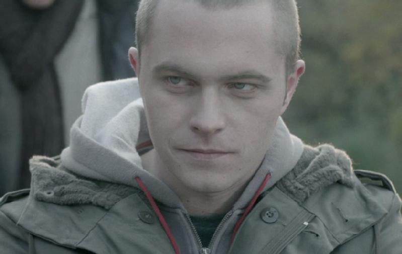 Młody mężczyzna z krótkimi włosami i poważnym wyrazem twarzy, ubrany w zieloną kurtkę z kapturem i szarą bluzę z kapturem pod spodem, stoi na zewnątrz w łagodnym, naturalnym świetle.