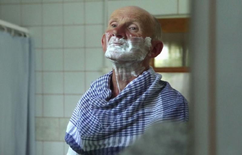 Starszy mężczyzna z kremem do golenia na twarzy stoi w łazience i patrzy w górę, mając na ramionach przewieszony niebiesko-biały ręcznik w kratkę.