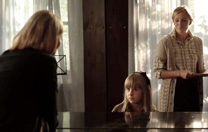 Młoda dziewczyna siedzi przy pianinie, patrząc na kobietę naprzeciwko niej, podczas gdy inna kobieta stoi w pobliżu, trzymając papiery. Miękkie światło dzienne sączy się przez cienkie firanki w tle.