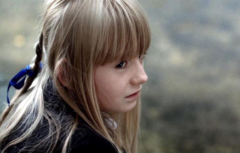 Młoda dziewczyna z długimi blond włosami i grzywką, ubrana w ciemny top, patrzy na bok. Jej włosy są związane niebieską wstążką, a ona sama wydaje się zamyślona lub zamyślona. Tło jest rozmyte i wyciszone.