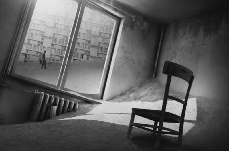 Pochylony, opuszczony pokój z jednym drewnianym krzesłem i piaskiem pokrywającym podłogę, promienie słońca wpadające przez duże okno; na zewnątrz ktoś przechodzi obok wysokich, jednolitych budynków mieszkalnych.