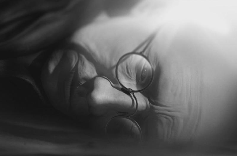 Starsza osoba w okrągłych okularach leży na boku w miękkim świetle, częściowo przesłoniętym przez cień, o spokojnym i zamyślonym wyrazie twarzy.