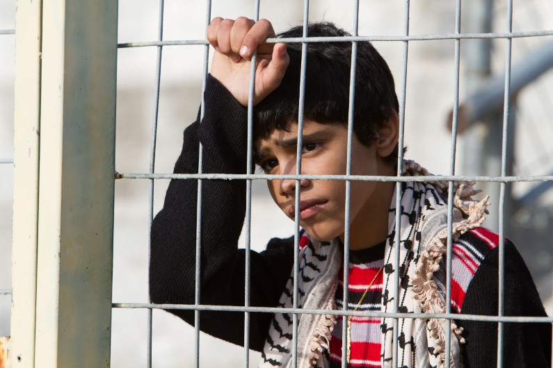 Młody chłopiec o ciemnych włosach i wzorzystym szaliku stoi za metalowym płotem, patrzy w dal z ponurym wyrazem twarzy, ręką ściskając kraty.