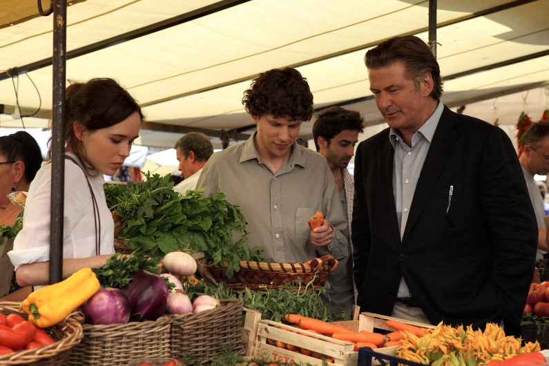 Trzy osoby stoją przy straganie na targu na świeżym powietrzu wypełnionym świeżymi warzywami, w tym marchewkami i zieleniną liściastą. Jedna osoba ogląda marchewkę, podczas gdy inni patrzą. Scena jest żywa i dobrze oświetlona pod baldachimem.