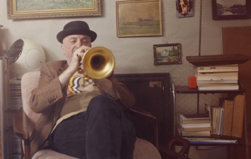Mężczyzna w kapeluszu i kraciastej marynarce siedzi w fotelu i gra na trąbce w przytulnym, zabytkowym pokoju, ozdobionym oprawionymi obrazami i stosami książek.