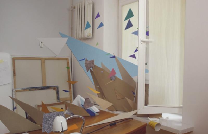 Zagracone biurko w małym pokoju, na którym porozrzucane są kolorowe, geometryczne kształty i wycinki z papieru, a także elementy unoszące się w powietrzu przy otwartym oknie, przez które wpada światło słoneczne.