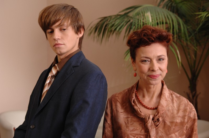 Młody mężczyzna w niebieskim garniturze i pasiastym krawacie stoi obok starszej kobiety w brązowej bluzce i czerwonym naszyjniku. Oboje stoją twarzą do kamery przed doniczkową rośliną, z neutralnymi wyrazami twarzy.