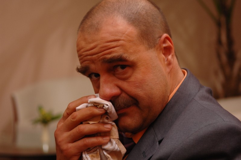 Mężczyzna w garniturze trzyma chusteczkę przy nosie i patrzy w kamerę z poważnym wyrazem twarzy. Tło jest delikatnie rozmyte, co sugeruje wnętrze z jasnymi ścianami i kilkoma roślinami.