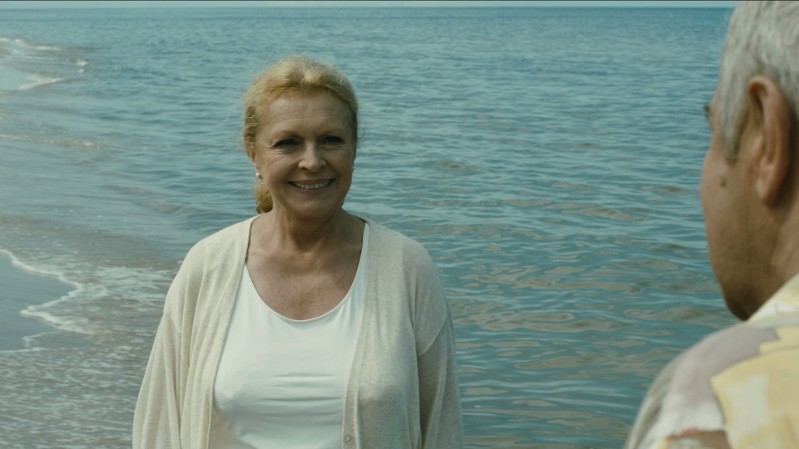 Kobieta o blond włosach, ubrana w białą bluzkę i lekki kardigan, stoi i uśmiecha się na plaży, blisko brzegu, zwrócona twarzą do starszego mężczyzny we wzorzystej koszuli, na tle oceanu.