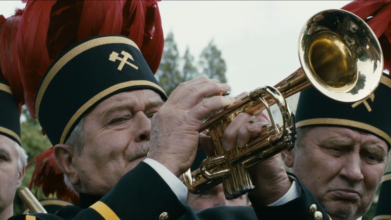 Mężczyzna w ciemnym mundurze i czapce z piórkiem gra na trąbce; obok niego stoi drugi mężczyzna w podobnym stroju. Obaj znajdują się na zewnątrz, w tle widać drzewa.