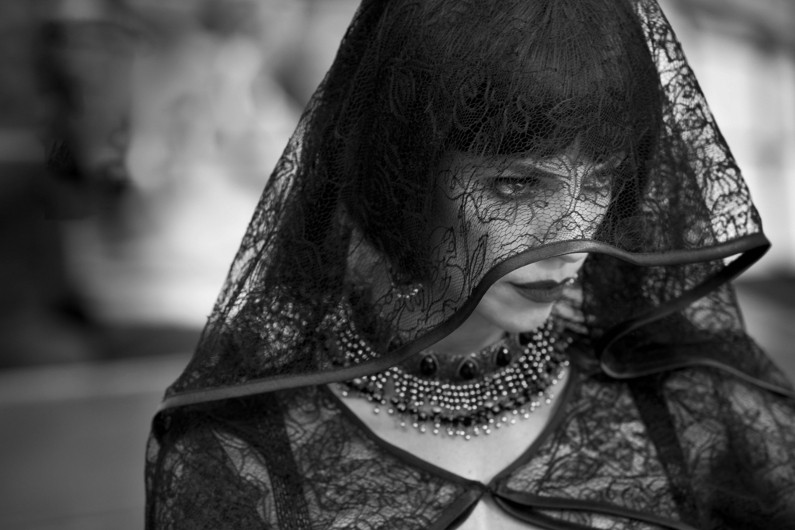 Kobieta ubrana w ciemny koronkowy welon i ozdobny naszyjnik z koralików spogląda w bok. Jej wyraz twarzy jest ponury, a obraz jest czarno-biały, co podkreśla fakturę koronki i nastrój.